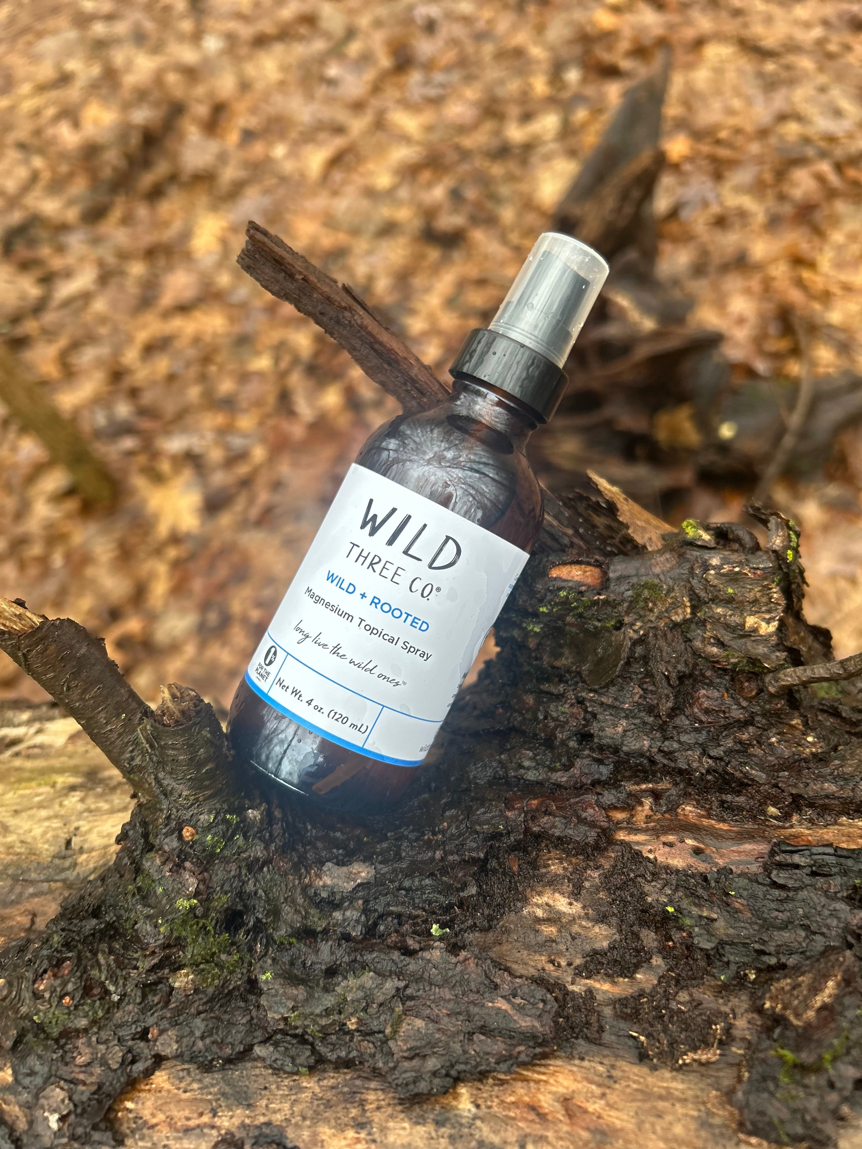 Wild Three Co. Magnesium Topical Spray is in an amber glass bottle styled with natural elements, highlighting a calm, nature inspired routine. The bottle is sitting on a log. Out of focus behind the bottle and log is a wooded area with trees and leaves on the ground. 