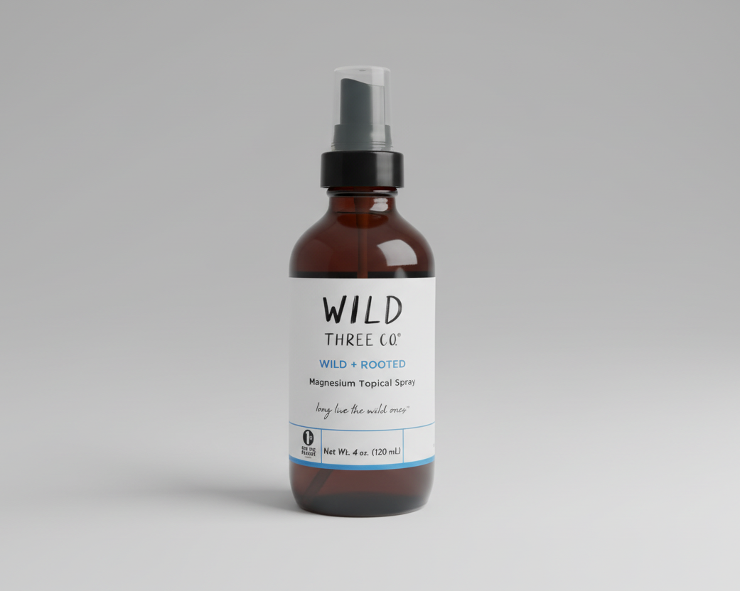 Wild Three Co. magnesium spray in amber glass bottle with black mist top and white minimalist label. The font on the label is black and blue. The bottle is sitting in front of a gray background. 