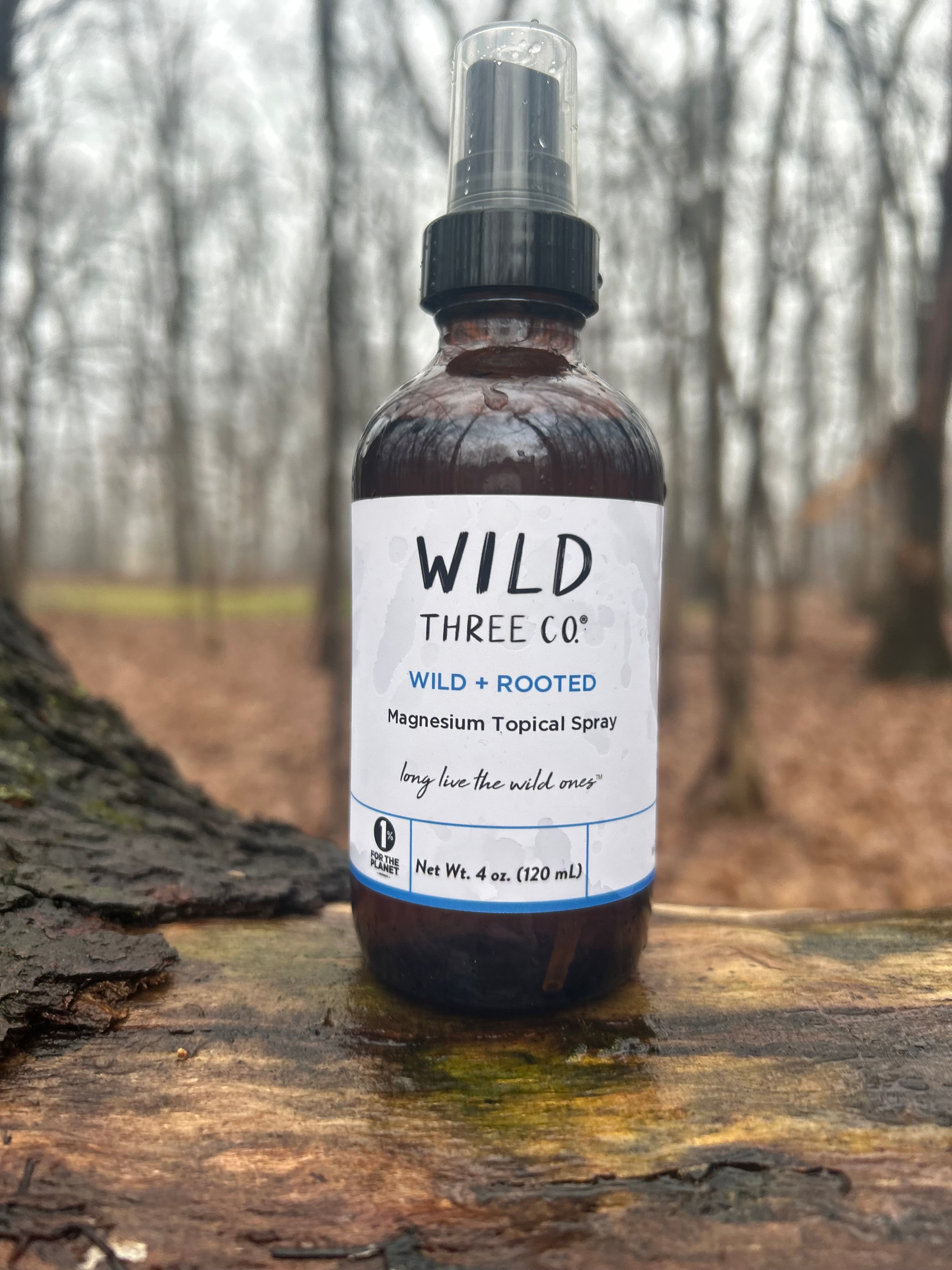 Wild Three Co. Magnesium Topical Spray is in an amber glass bottle styled with natural elements, highlighting a calm, nature inspired routine. The bottle is sitting on a log. Out of focus behind the bottle and log is a wooded area with trees and leaves on the ground. 