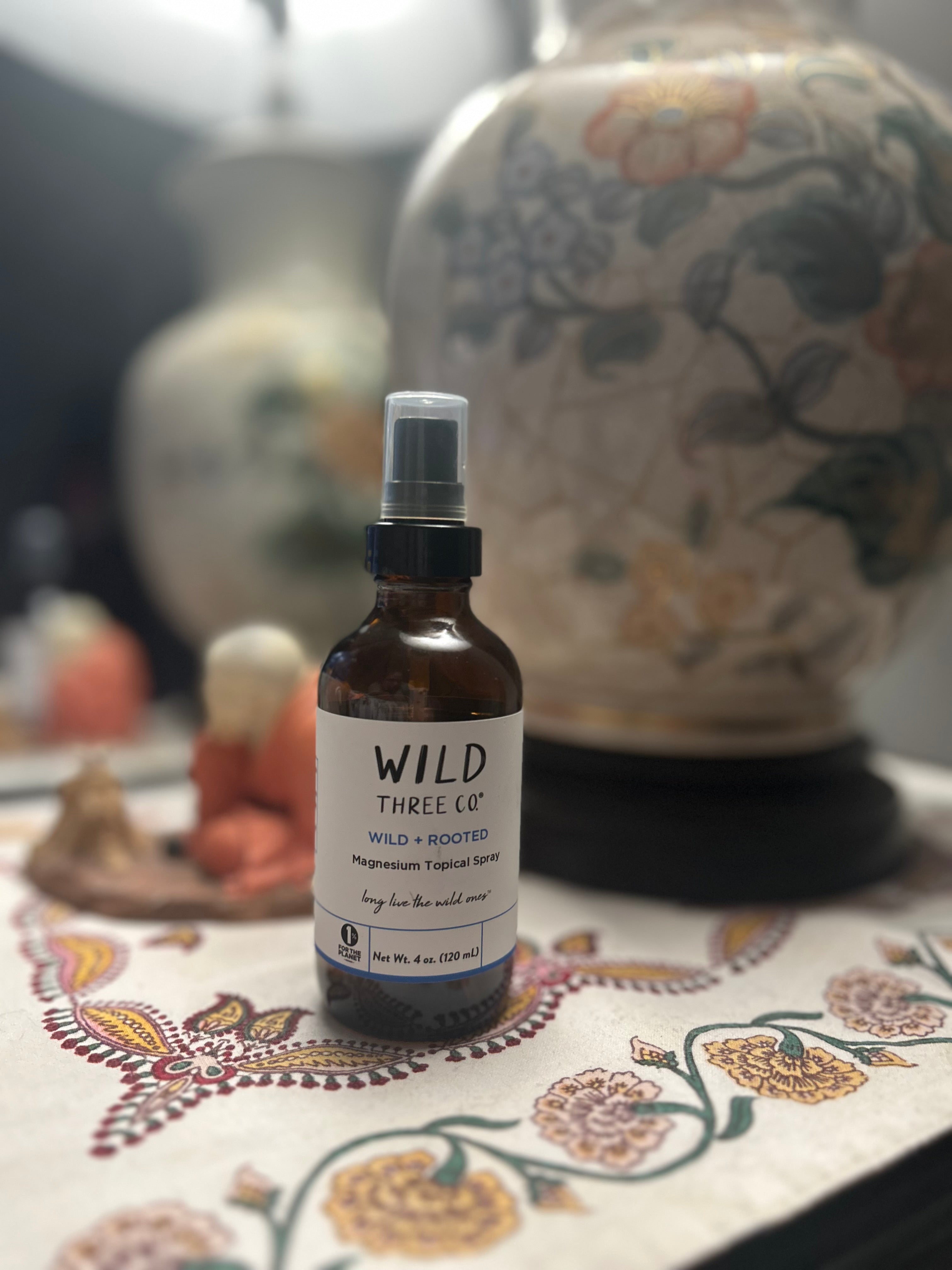 Wild Three Co. magnesium topical spray bottle is sitting on a bedside nightstand. The bottle is in focus. Out of focus behind the bottle, is an ornate lamp, a small monk statue, a mirror. The bottle, lamp and monk statue are sitting on a multicolored table runner. 