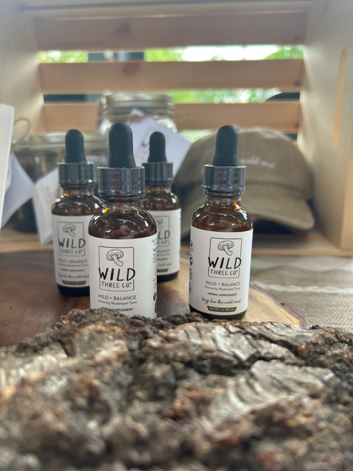 4 bottles of 'Wild + Balance' products displayed on a wooden surface with a piece of bark sitting in front of the bottles. In the background are items that are blurry but you can tell that there is a hat and glass jars and a wooden crate. The sun is shinning through the crate. The amber tincture bottles have a white label with the logo and product name on the front along with the tagline 