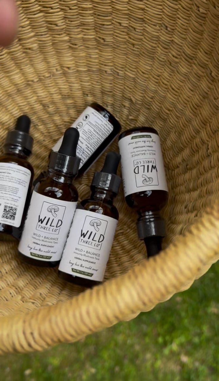 Hand holding a woven basket filled with 5 amber tincture bottles with white labels. The bottles are randomly sitting in the woven basket haphazardly. The bottles are of Wild+Balance mushroom immunity tonic. There is a grassy background. 