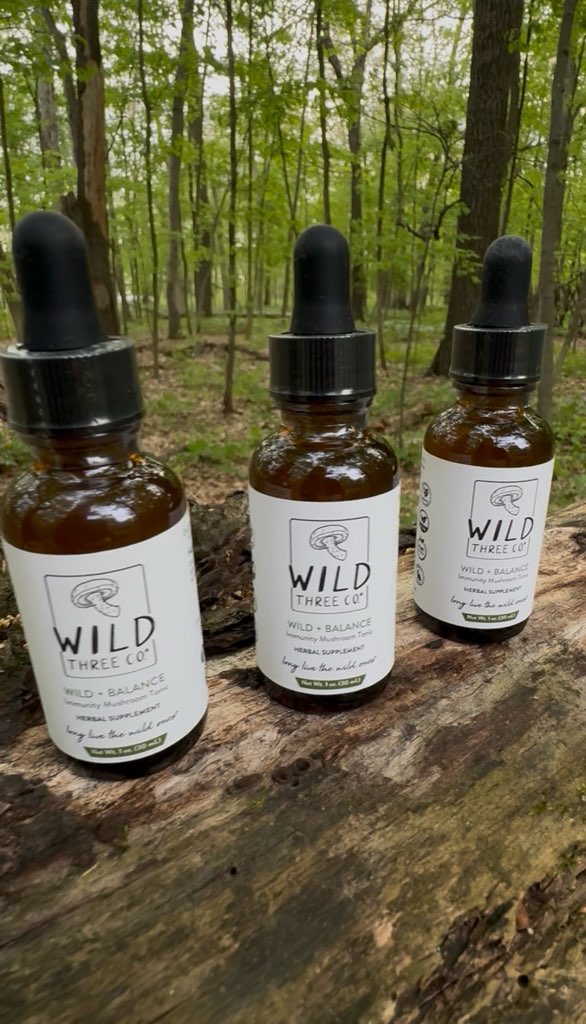 Three bottles of 'Wild + Balance' products on a wooden log with a forest background. The amber tincture bottles have a white label with the logo and product name on the front along with the tagline 